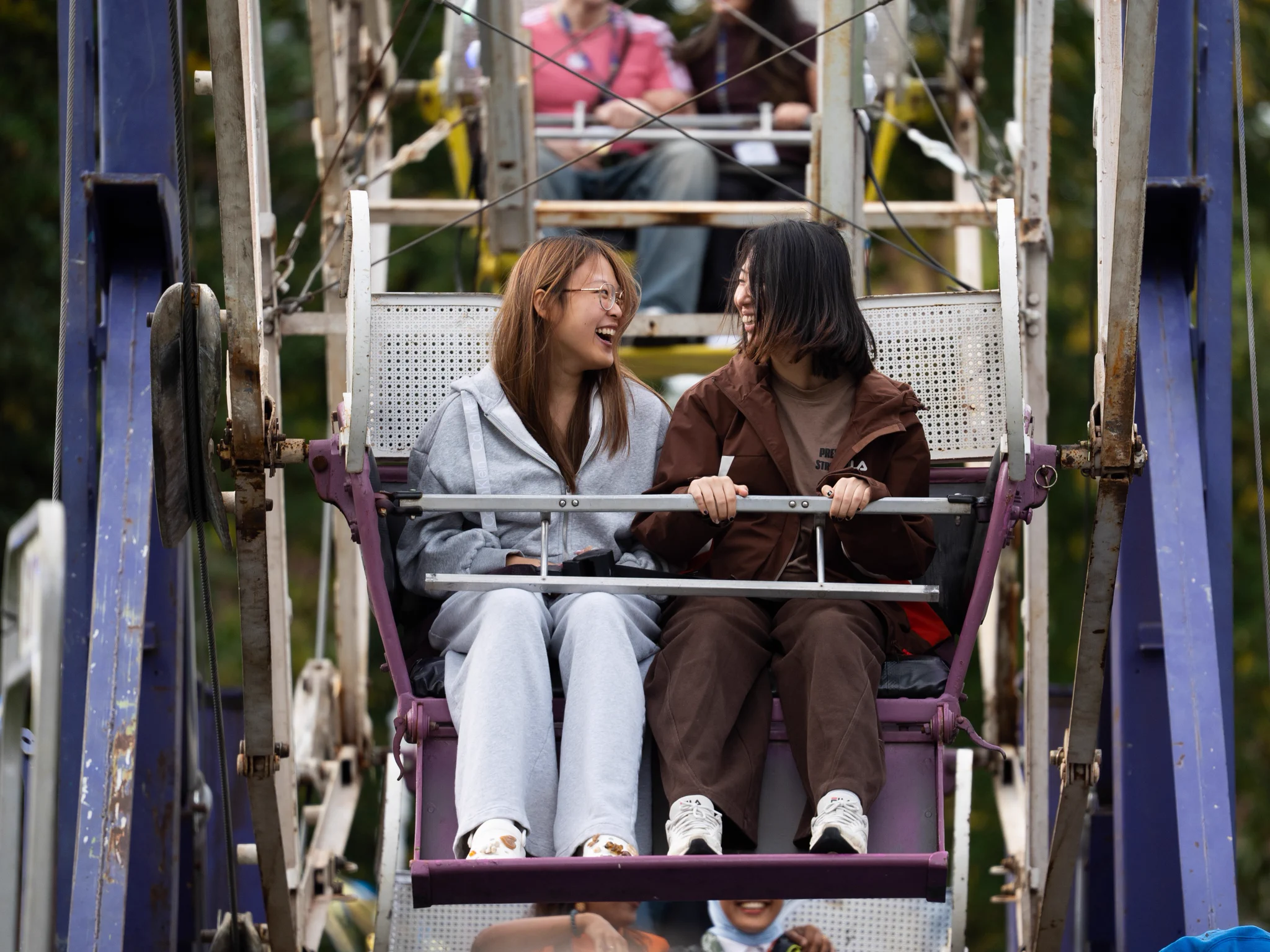 Two people are sitting together on a Ferris wheel, smiling at each other. They are enjoying a ride outdoors, surrounded by greenery.