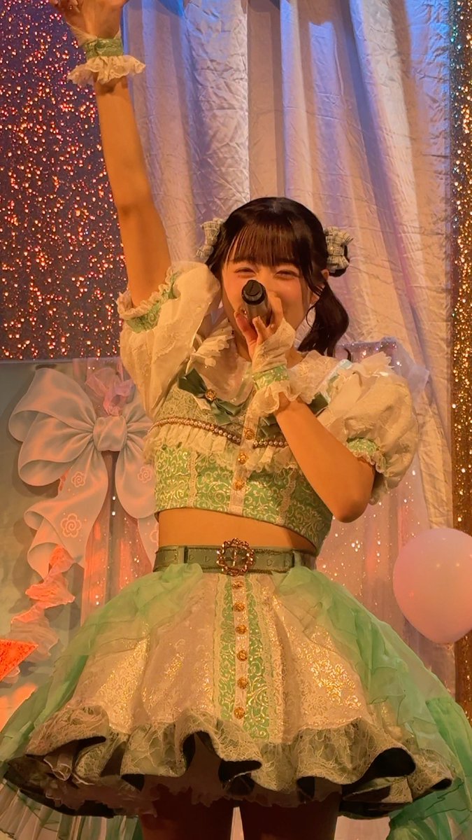 A young woman in a green and white frilled idol costume sings enthusiastically into a microphone on a brightly decorated stage with pastel curtains and large bows.