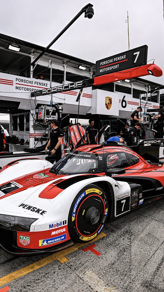 A Porsche Penske Motorsport #7 race car is parked in its pit garage with crew members working around it, featuring sponsor logos including Penske, TAG Heuer, Mobil 1, Motul, and Michelin.