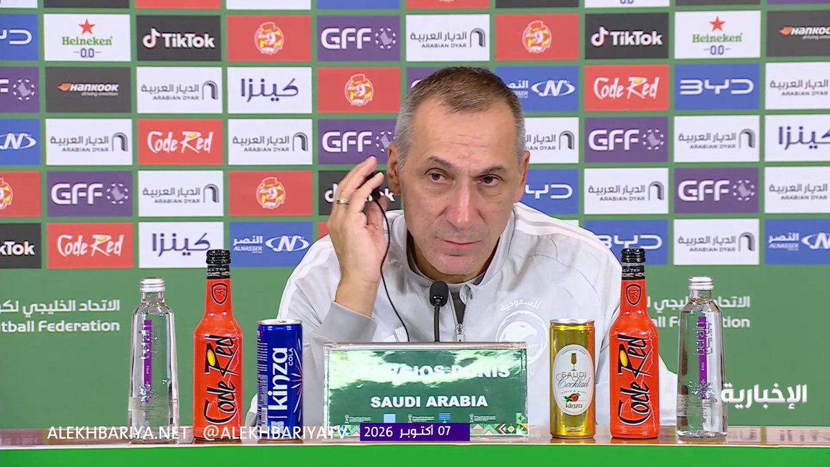 Saudi Arabia's national team coach Georgios Donis speaks at a press conference, seated before a backdrop of sponsor logos including Heineken, TikTok, Hankook, and GFF, with a placard reading "SAUDI ARABIA" and various beverages (Code Red, Kinza, and water bottles) arranged on the table before him. The image visually accompanies his statement about focusing on team development and defensive performance, as he gestures thoughtfully while addressing the media.