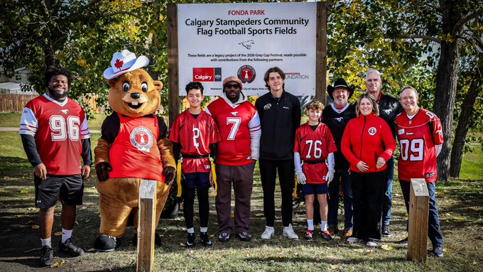 Group photo of people wearing Calgary Stampeders CFL jerseys (numbers 99, 7, 76, 09) and a bear mascot in a cowboy hat, posed in front of a sign reading 'FONDA PARK Calgary Stampeders Community Flag Football Sports Fields' with partner logos.