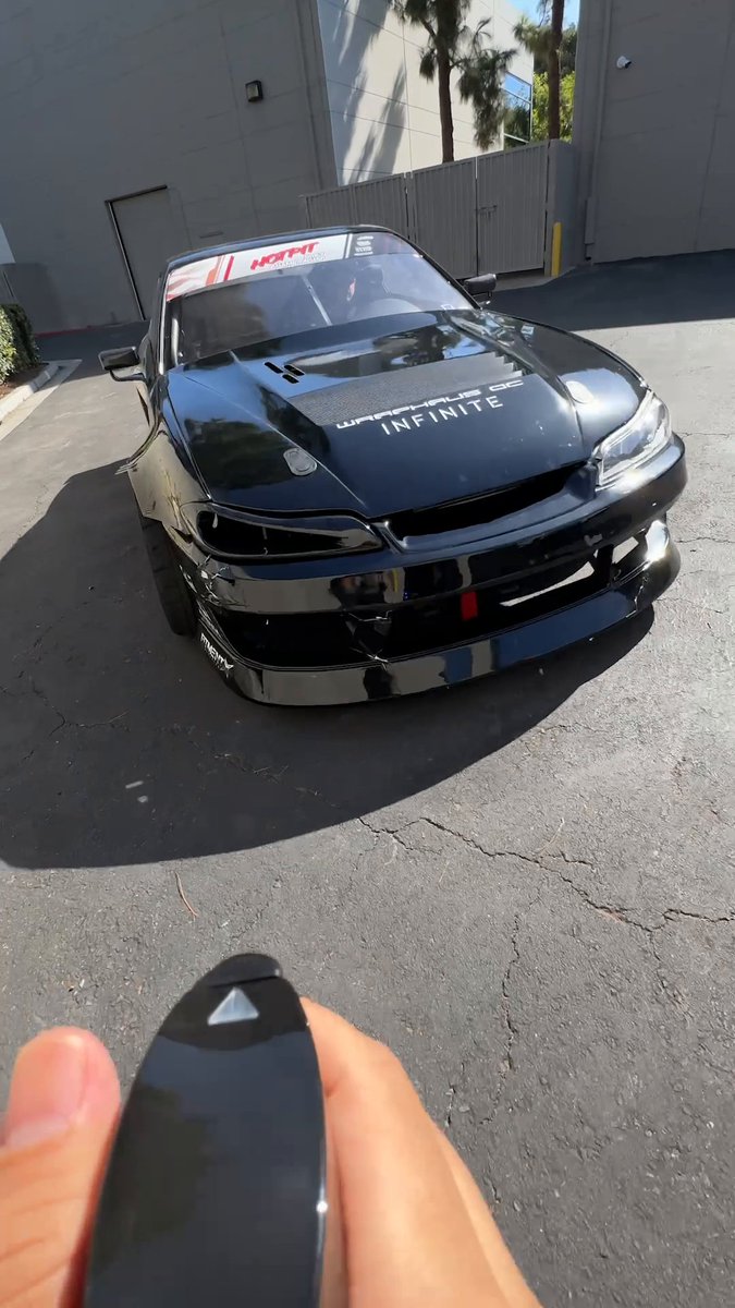 A black modified car sits in a parking lot, displaying 'INFINITE' and 'WARSPLUS DC' decals on the hood along with a 'HGMT' windshield banner, while a hand holds a car key fob in the foreground.