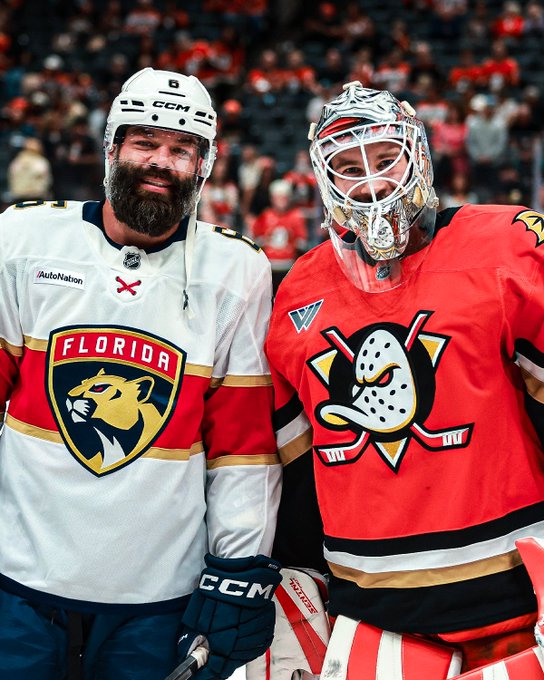 Two hockey players pose together on the ice, with a bearded player in a white Florida Panthers jersey (featuring "FLORIDA" and "AutoNation" text and a #6 helmet) standing next to a goalie wearing an Anaheim Ducks red jersey with the iconic duck goalie mask and number "DC 87" displayed. The image illustrates the post's caption about a Florida Panthers player reuniting with former teammate Lukas Dostal, who now plays for the Anaheim Ducks.