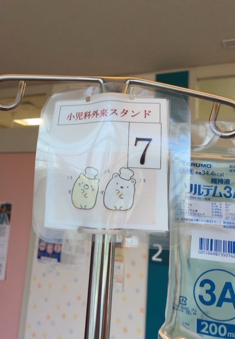 A close-up of an IV drip chamber at a Japanese medical facility, with a tag reading '小児科外来スタンド 7' featuring small bear drawings, alongside a 3A 200ml saline bag.