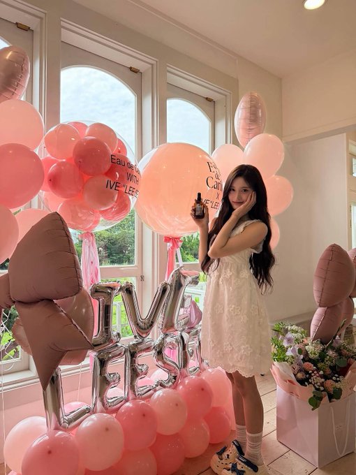 A young woman in a white dress posing in a brightly decorated room filled with pink and brown balloons, large 'BIRTHDAY' foil letters, and a bouquet of flowers.