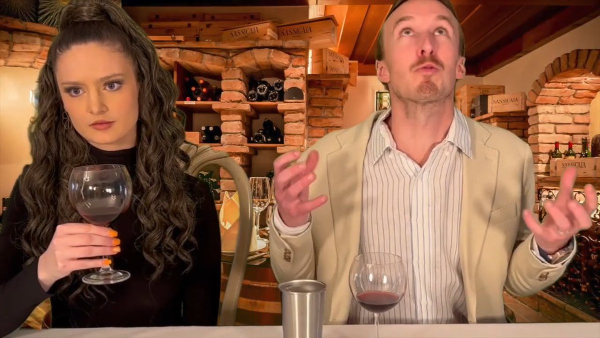 A woman holding a glass of red wine and a mustached man with raised hands and pursed lips stand in a wine cellar with brick alcoves and wooden crates labeled 'SASSICAIA' displayed on the shelves behind them.