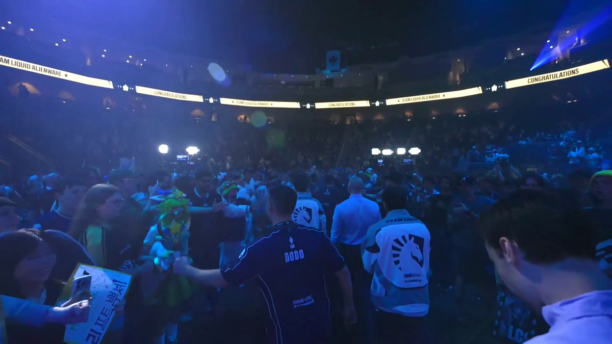 Team Liquid players and fans celebrate their Worlds championship victory in an arena lit by blue spotlights, with LED screens displaying "CONGRATULATIONS" and "TEAM LIQUID ALIENWARE" overhead. The crowd, including players wearing jerseys marked "DODO" and the recognizable Team Liquid horse logo, swarms the stage area in jubilation.