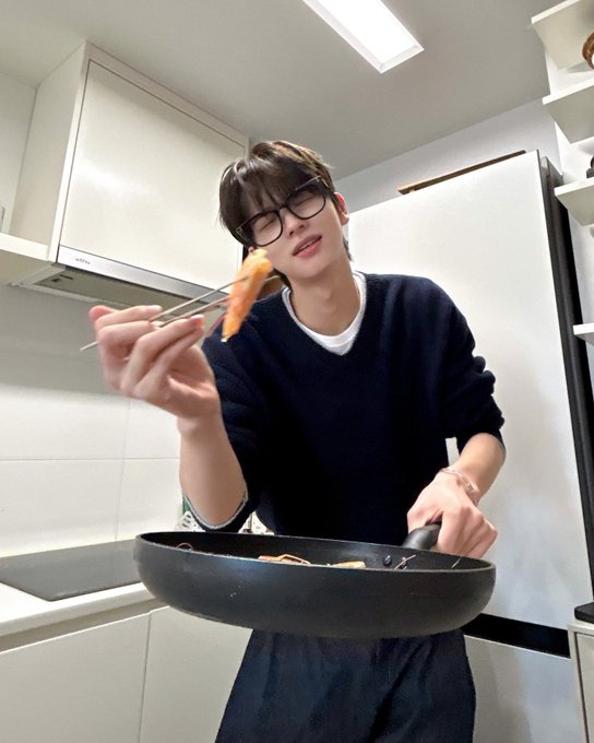 A young man with glasses, identified as SHINYU from TWS, stands in a kitchen holding a black pan and chopsticks with a piece of food, appearing to enjoy a meal he's cooking. The post ties into the image by suggesting his cooking is so good he'll never need public transportation again, matching his cheerful expression as he offers the food toward the camera.