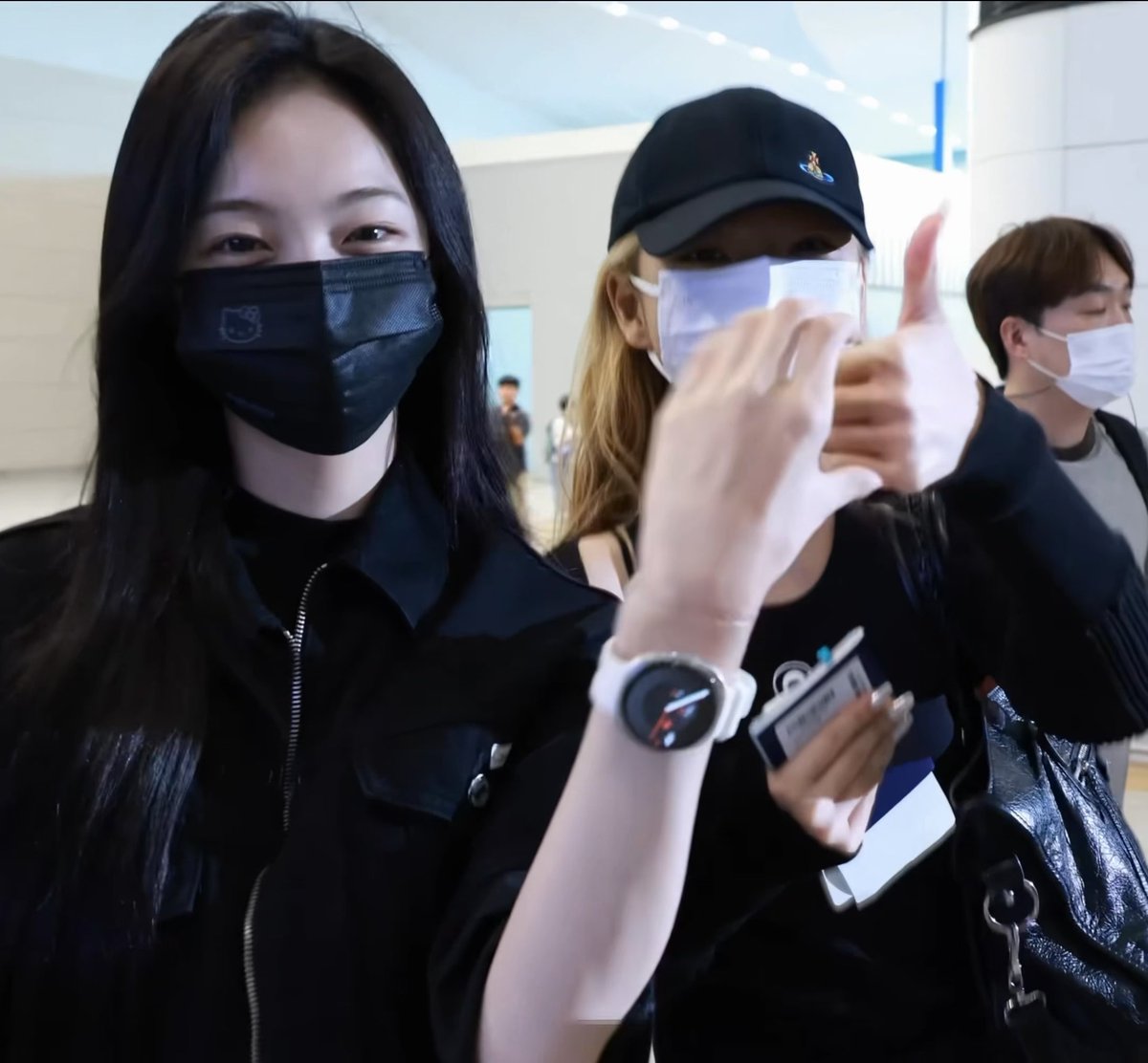 Two young women at what appears to be an airport both give a thumbs-up; one wears a black Hello Kitty mask while the other wears a black cap and white mask.