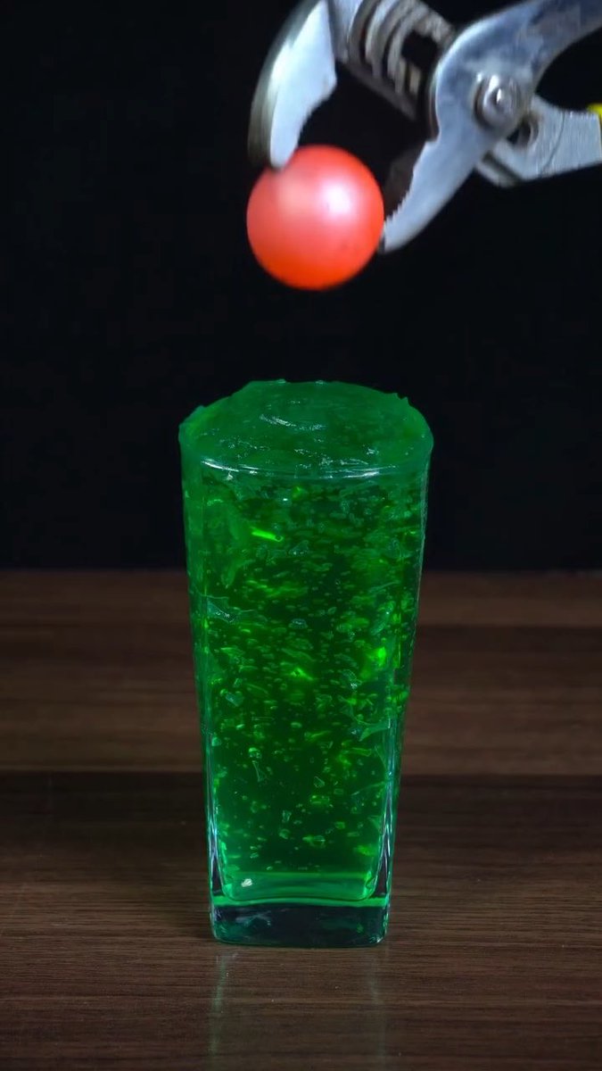 A still image of a red ping pong ball being held by a metallic claw mechanism above a tall glass filled with a bright green carbonated beverage on a wooden surface against a black background.