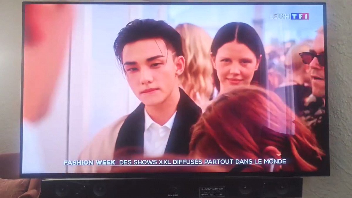 A television screen displays TF1's live broadcast of Fashion Week, showing the French news ticker "FASHION WEEK DES SHOWS XXL DIFFUSÉS PARTOUT DANS LE MONDE," with a young man (Hyunjin) featured prominently, presumably from his appearance at the Dior Summer 2027 show in Paris as noted in the accompanying caption.