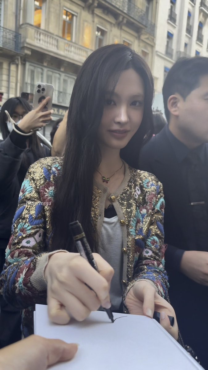 A woman wearing an ornate gold and floral-embroidered jacket signs a document with a pen, with 'lisasempi' text visible on the image.