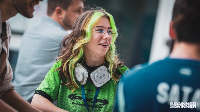 A woman with neon green-streaked hair wearing glasses, a green jersey, and headphones around her neck, smiling in conversation at what appears to be an esports event with 'BGSKINNER' watermark.