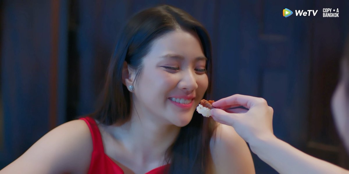 A smiling young woman in a red top is being offered a bite of food (rice with a topping) by another person's hand, with a "WeTV" logo and "COPY A BANGKOK" text in the upper right corner.