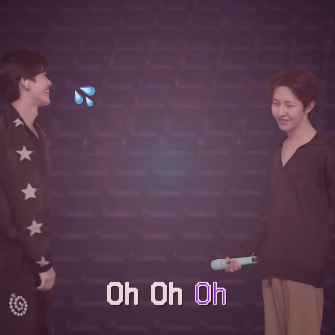 Two Asian men stand on a stage against a dark brick wall background, smiling at each other, with one holding a light blue microphone and pixelated text reading 'Oh Oh Oh' and a sweat drop emoji overlaid.