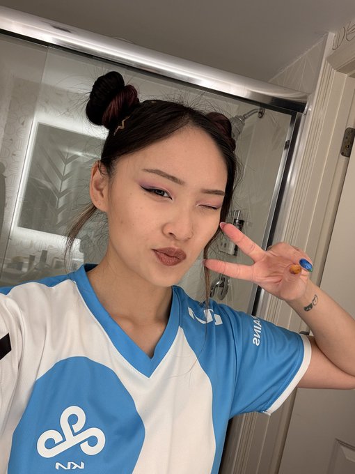 A selfie of a young woman with space buns hairstyle, pink eyeshadow, and a blue and white Cloud9 esports jersey, making a peace sign near a bathroom mirror.