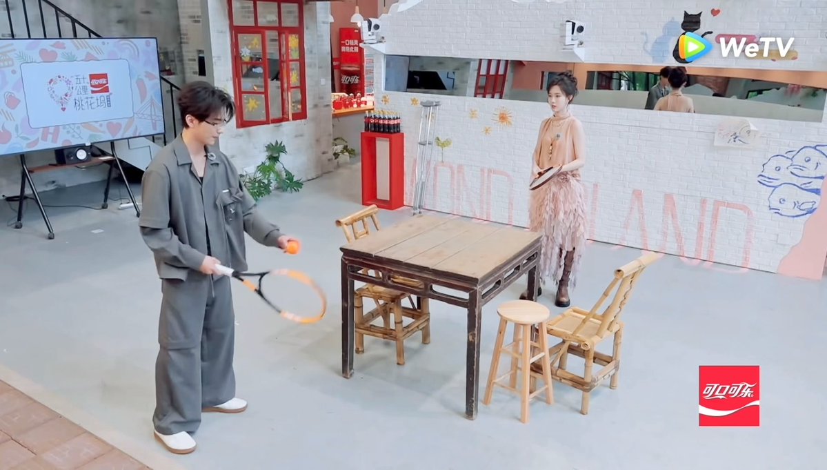 A man in a gray outfit holds a tennis racket and ball while standing across from a woman in a feathered skirt at a wooden table on a set branded with 'WeTV', '桃花坞' (Peach Blossom Land), and Coca-Cola logos.