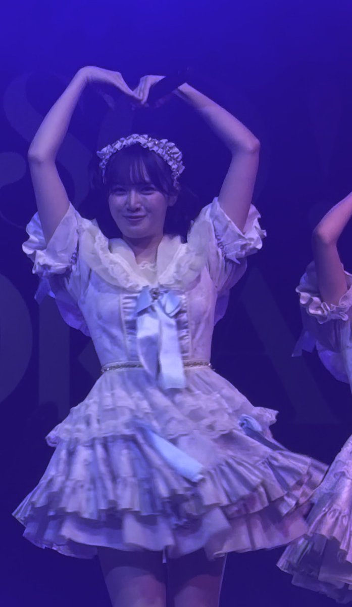 A young woman wearing a frilly light purple dress with a headband smiles and forms a heart with her hands above her head during a stage performance.