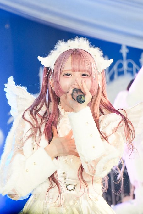 A performer with long pink hair and a white angel-themed costume sings into a microphone on stage, wearing a fluffy halo headband.