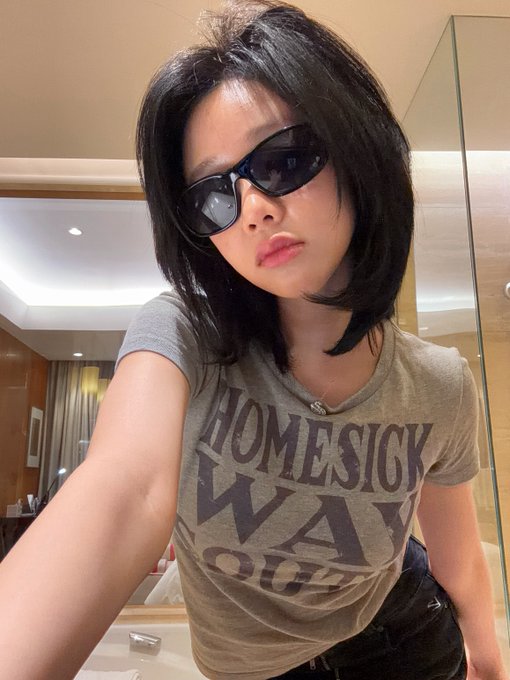 A young woman with dark hair takes a selfie wearing black sunglasses and a grey t-shirt with the text 'HOMESICK WACO' printed on it.