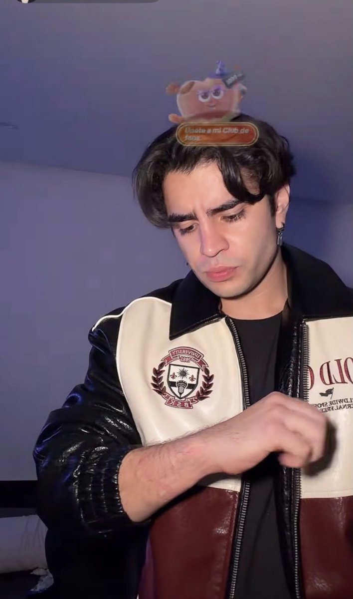 A young man with dark hair looks downward while wearing a two-toned leather jacket; an animated sticker with Arabic text and a small animal character is superimposed on his head.