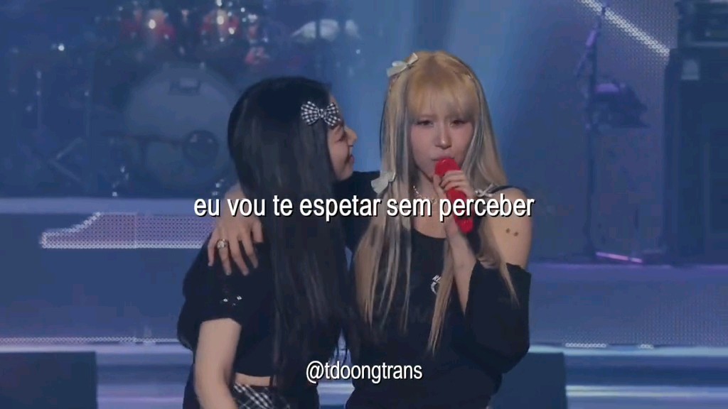 Two women stand on stage; one holds a red microphone, and overlaid text reads "eu vou te esp te esp te esp etar sem perceber" with a watermark "@tdoongtrans" below.