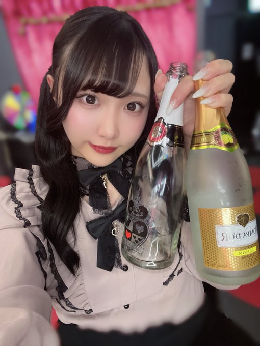 A young woman with dark hair in a gothic-style blouse holds up two bottles, one featuring a Mickey Mouse sticker, in what appears to be a bar or party setting.