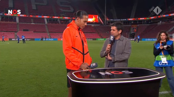 A TV interview on the NOS network taking place at the Johan Cruyff Arena stadium, where a reporter holds a microphone interviewing a man in an orange jacket with a photographer in the background.