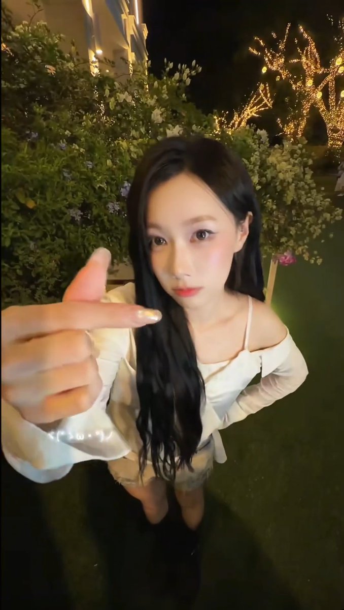 A young woman with long black hair, wearing a white off-shoulder top and beige skirt, pointing at the camera while standing outdoors at night near illuminated decorative trees.
