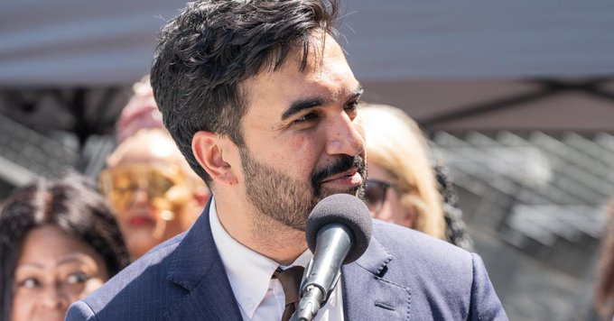 A close-up photo of a bearded man in a dark blue suit speaking into a microphone at what appears to be an outdoor event.