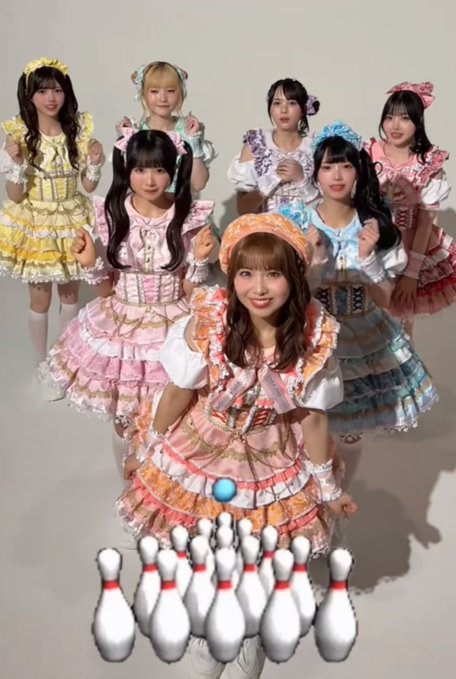 Group of Japanese performers in colorful frilly dresses posing together, with an edited bowling pin arrangement overlaid at the foreground.