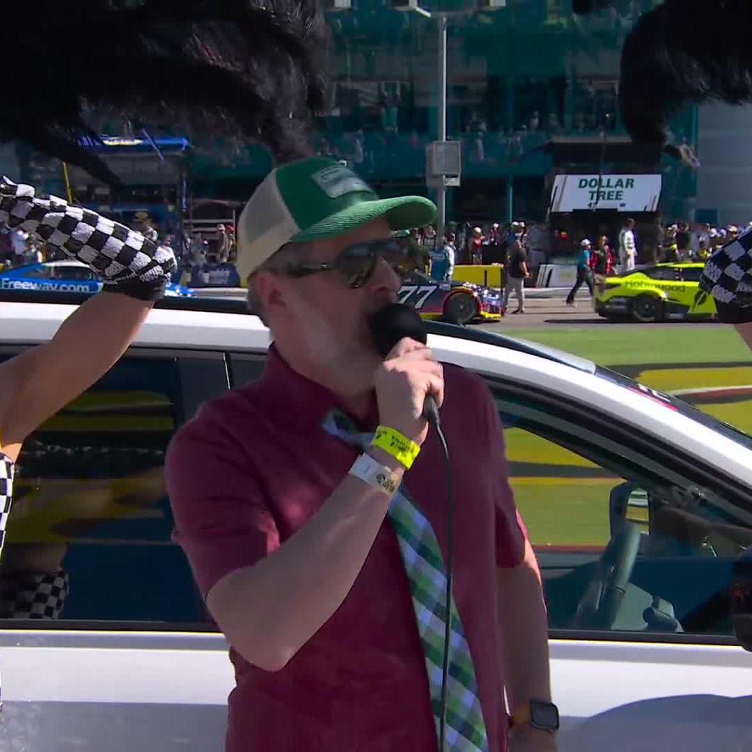 A man in a maroon polo shirt, green and white cap, sunglasses, and a plaid tie speaks into a microphone while standing in front of a white SUV, with checkered flags framing the shot and race cars (#77) visible in the background near a Dollar Tree sign. The image aligns with the post's description of Brendan Hunt giving the command to fire engines at Las Vegas Motor Speedway before a race.