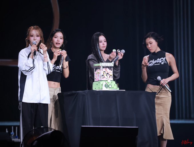 Four young women on stage holding microphones around a decorated cake, appearing to celebrate a fan meeting or idol event.