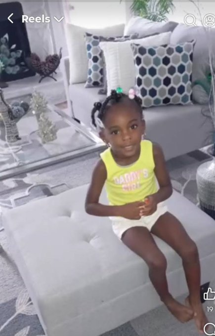 A young girl sitting on a gray ottoman in a living room, wearing a yellow tank top that says 'daddy's girl' and white shorts, with her hair styled in small buns.