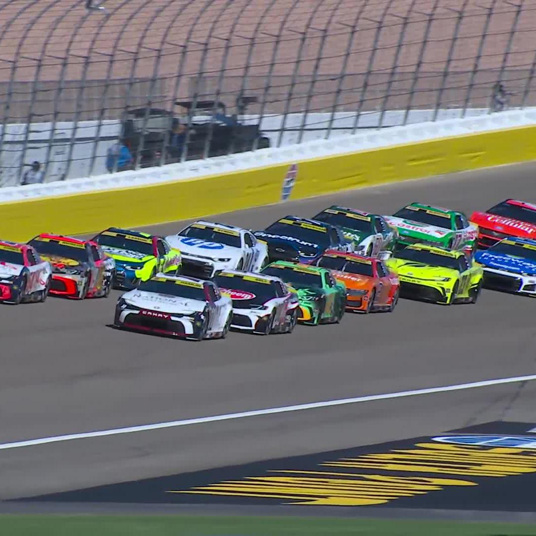 A pack of NASCAR stock cars, including ones sponsored by HP, Grainger, and Camping World, race in tight formation during the Sin City Showdown at LV Motor Speedway. The drivers are bunched together in multiple rows, battling for position as they speed down the track with the catch fence visible in the background.