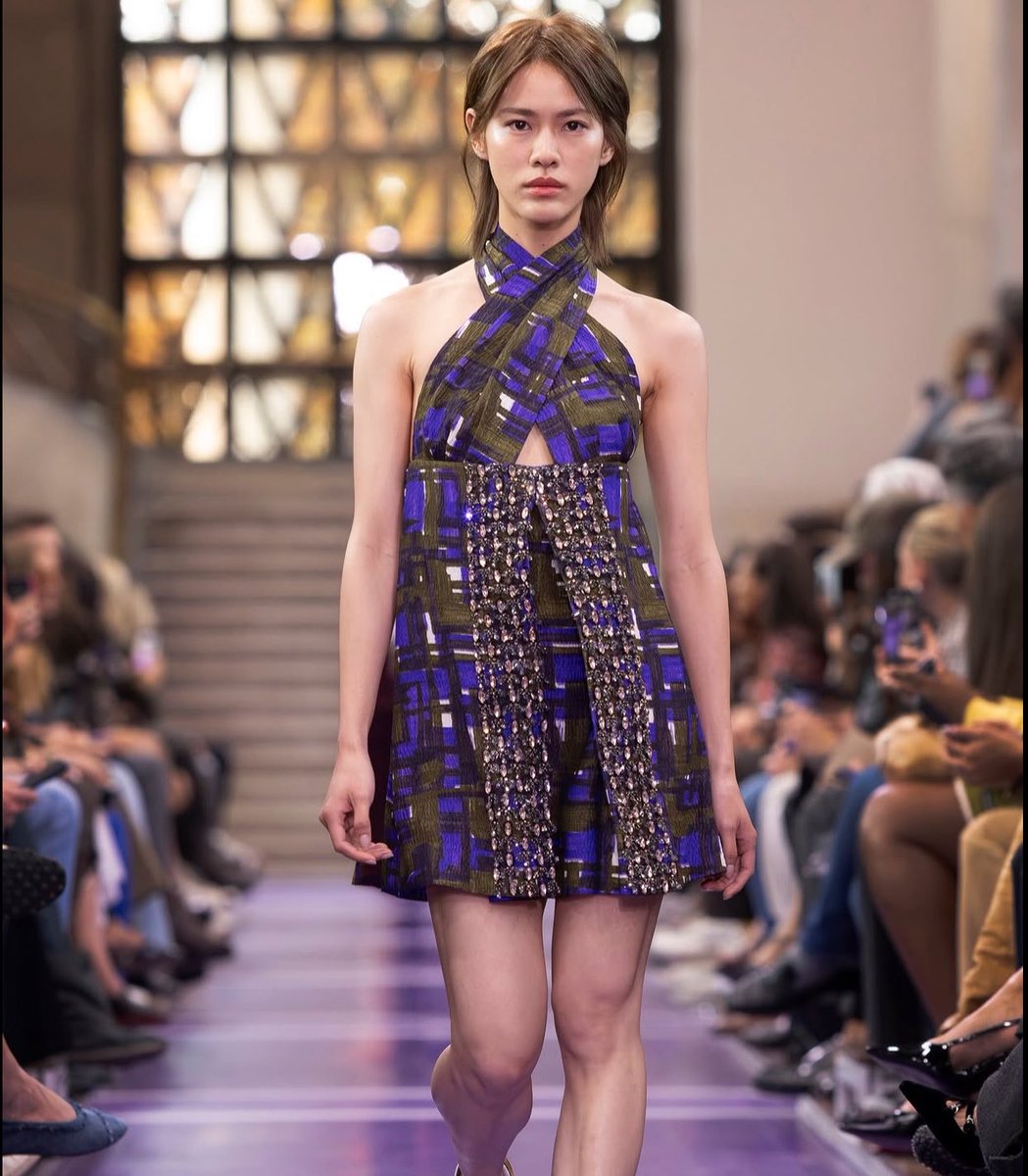 A model walks a runway wearing a blue, green, and white patterned halter-neck mini dress featuring crystal-embellished detailing, with an audience visible in the background.