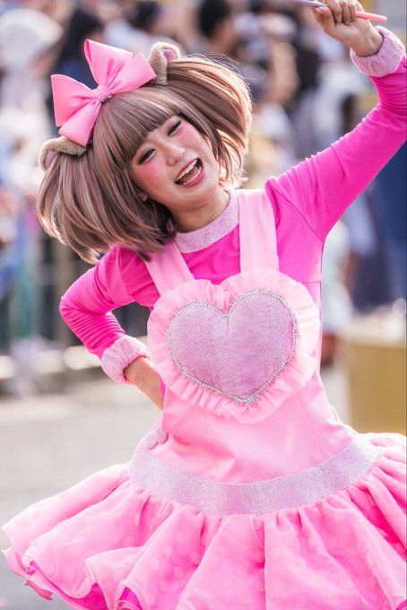 A performer in a bright pink costume with a heart on the front, pink bow, and pigtails poses with her hand on her hip at what appears to be a parade.
