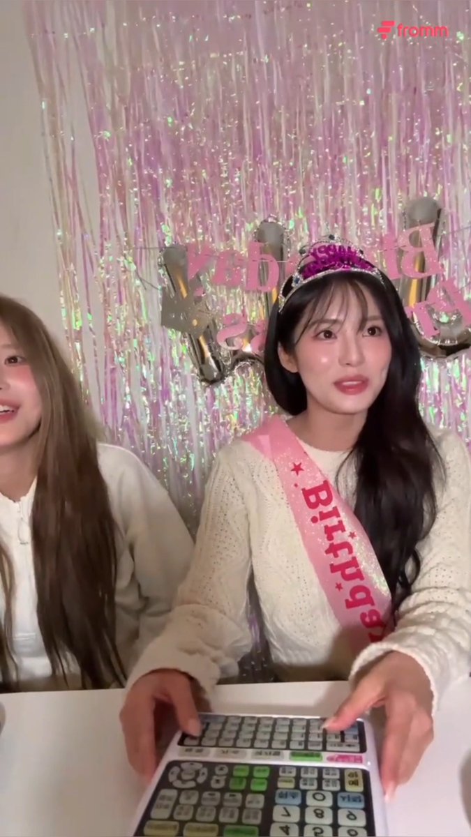 A woman wearing a "Birthday" sash and tiara sits at a table in front of a "Happy Birthday" banner, with another person partially visible beside her.