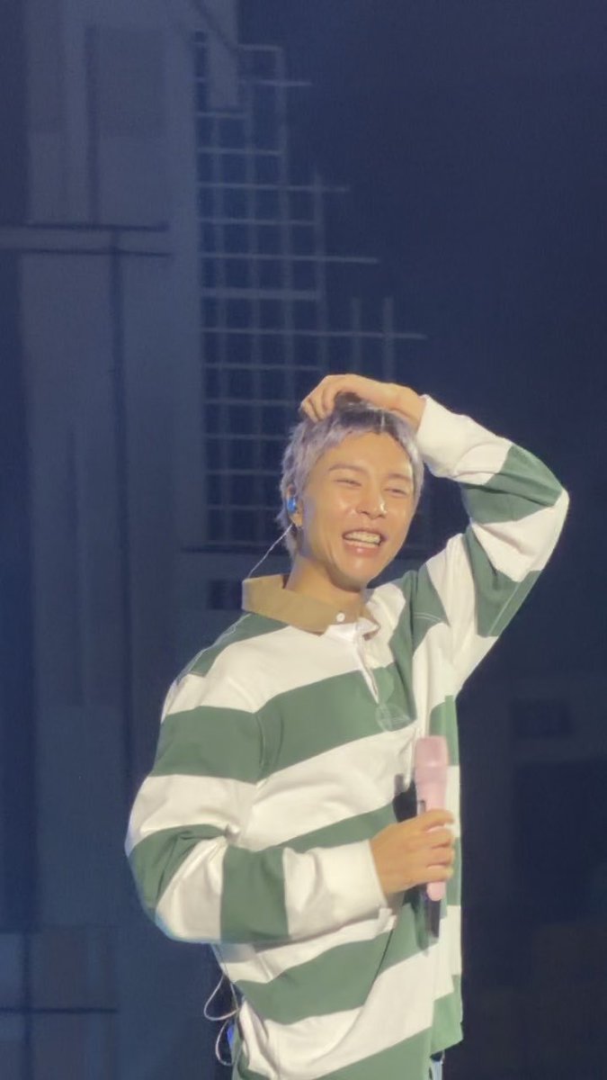 A performer with light purple hair, wearing a green and white striped shirt, smiles and runs a hand through their hair while holding a pink microphone on stage.