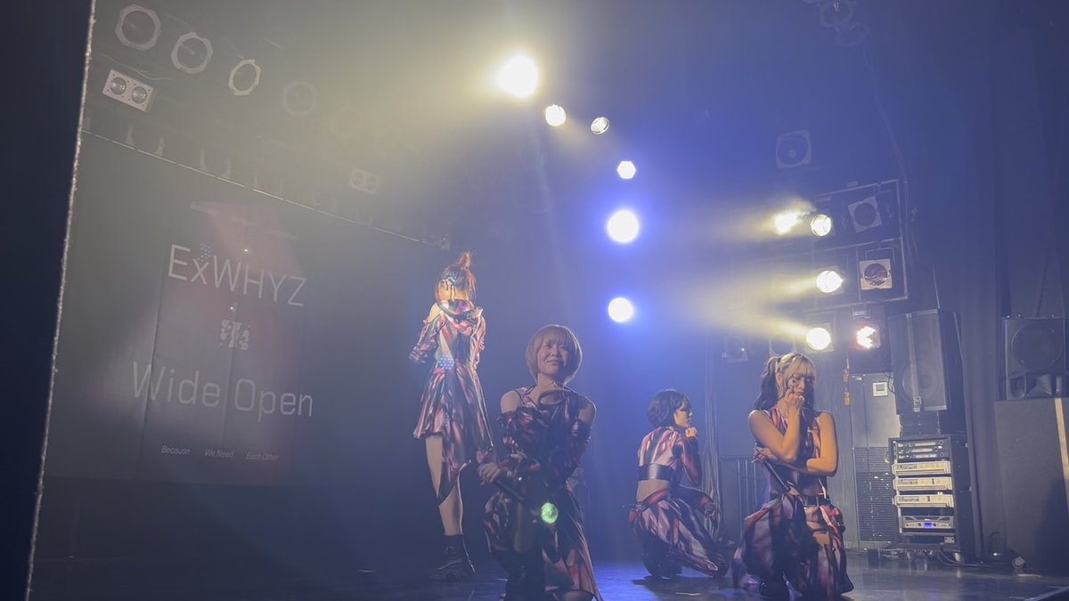 Four performers in coordinated patterned outfits pose on a stage with blue lighting; a banner in the background reads 'ExWHYZ Wide Open Because We Need Each Other.'