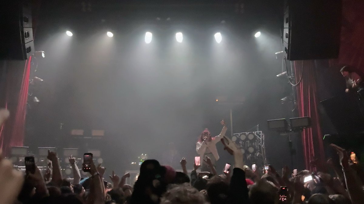 A concert scene viewed from the audience, showing a performer on stage under bright spotlights with fans raising phones and hands.