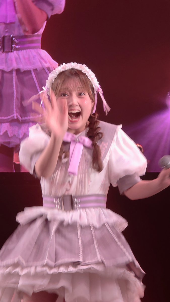 A young performer in a frilly white and purple maid-style dress with a matching headband smiles enthusiastically and waves to the audience while holding a microphone on stage.