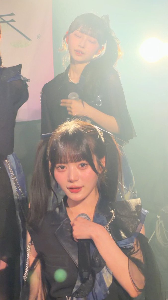 A concert photo showing two young female performers; the one in the foreground wears a black costume with pigtails and holds a microphone, while the one behind her in a dark shirt holds a microphone with eyes closed.