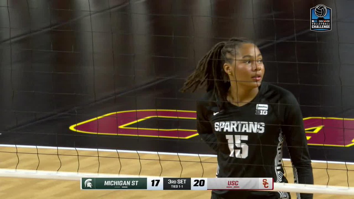 A Michigan State Spartans volleyball player (#15) in black stands at the net during a match against USC, with the scoreboard showing Michigan State 17, USC 20 in the 3rd set.