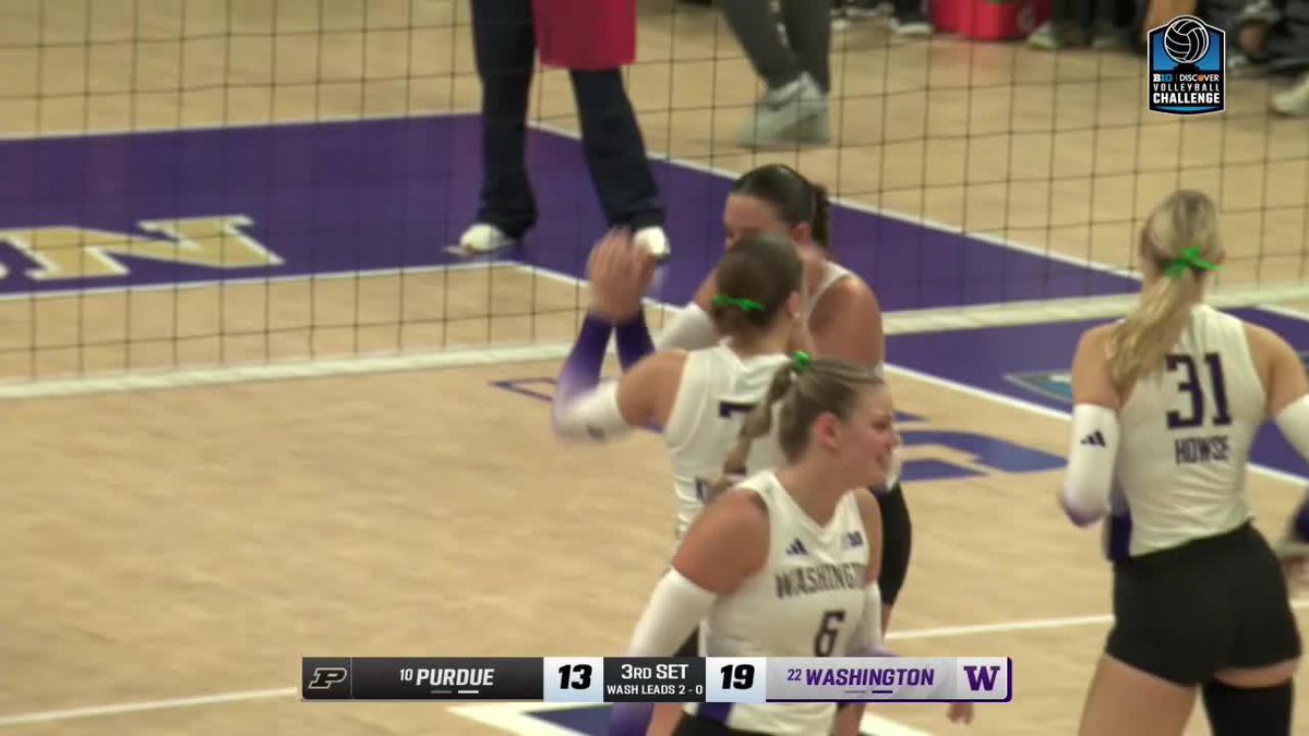 Washington volleyball players in white jerseys celebrate during a match against #10 Purdue, with the scoreboard showing Washington leading 19-13 in the 3rd set.