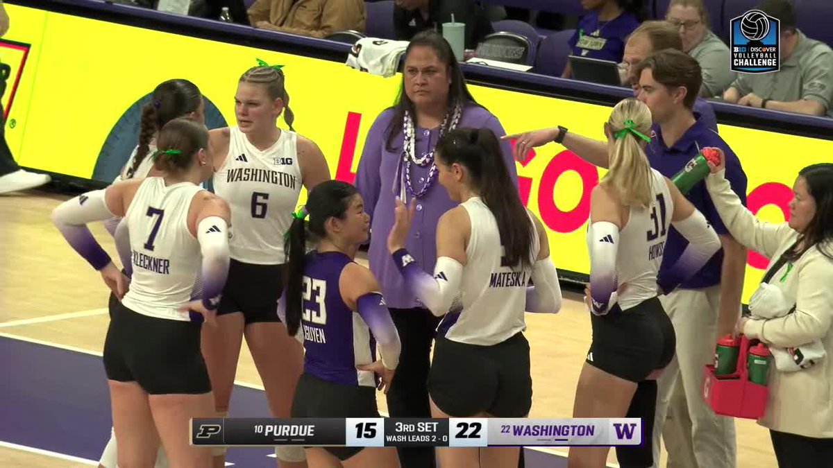 Members of the University of Washington volleyball team, wearing white jerseys with purple accents, huddling on the sideline during a Big Ten matchup; the scoreboard shows Purdue trailing Washington 15-22 in the 3rd set, with Washington leading 2-0.