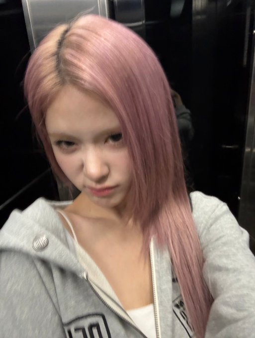 A selfie of a young woman with long pink hair wearing a gray hoodie, posing with a neutral expression against a dark background.