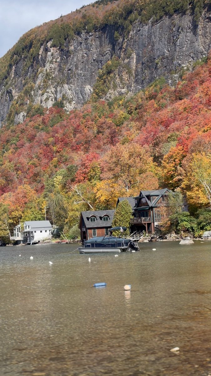 A serene autumn scene showing colorful fall foliage covering a rocky mountain, with a wooden house, a boathouse, and a pontoon boat on a calm lake.