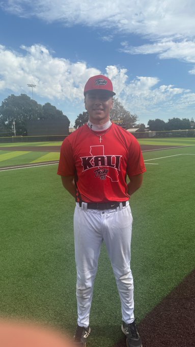 A young man in a red 'KBHL' baseball jersey, white pants, and matching cap stands on a baseball field under a partly cloudy sky.
