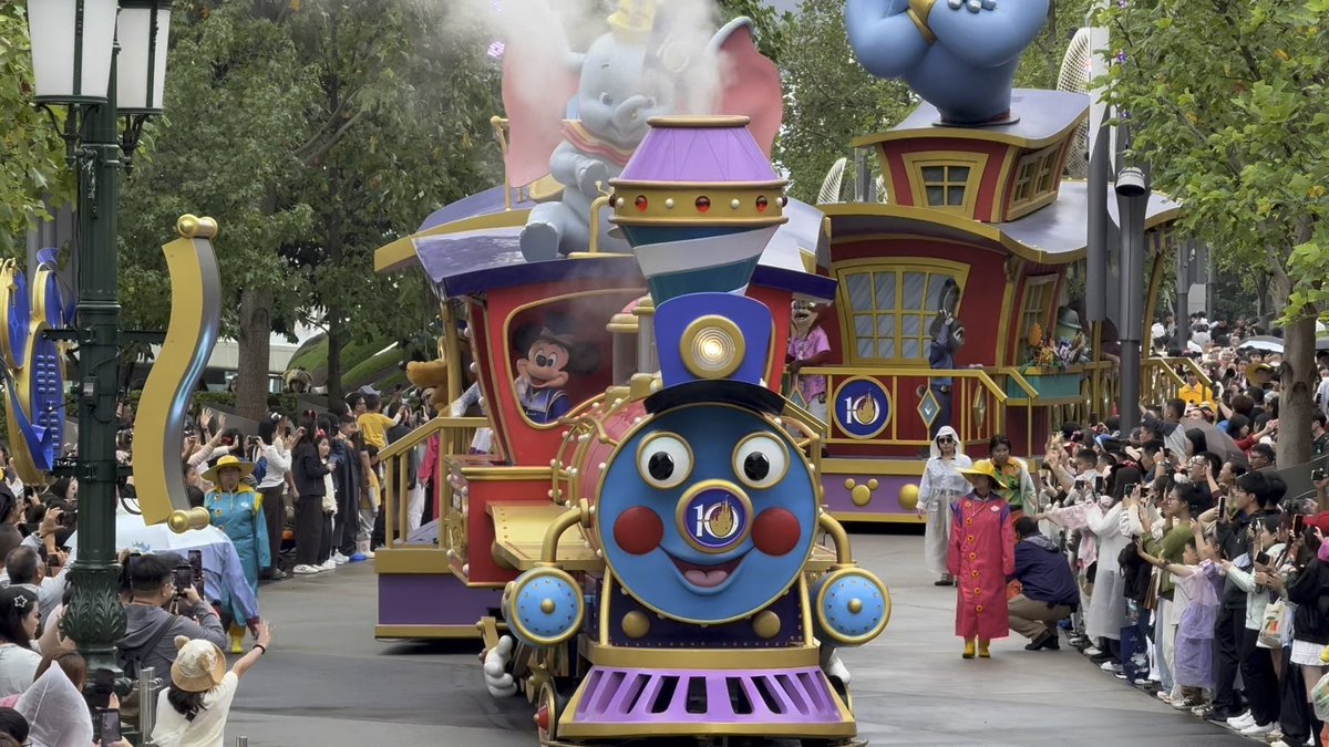 A Disney parade features a colorful train float with Mickey Mouse and Dumbo, accompanied by cast members and surrounded by cheering crowds.